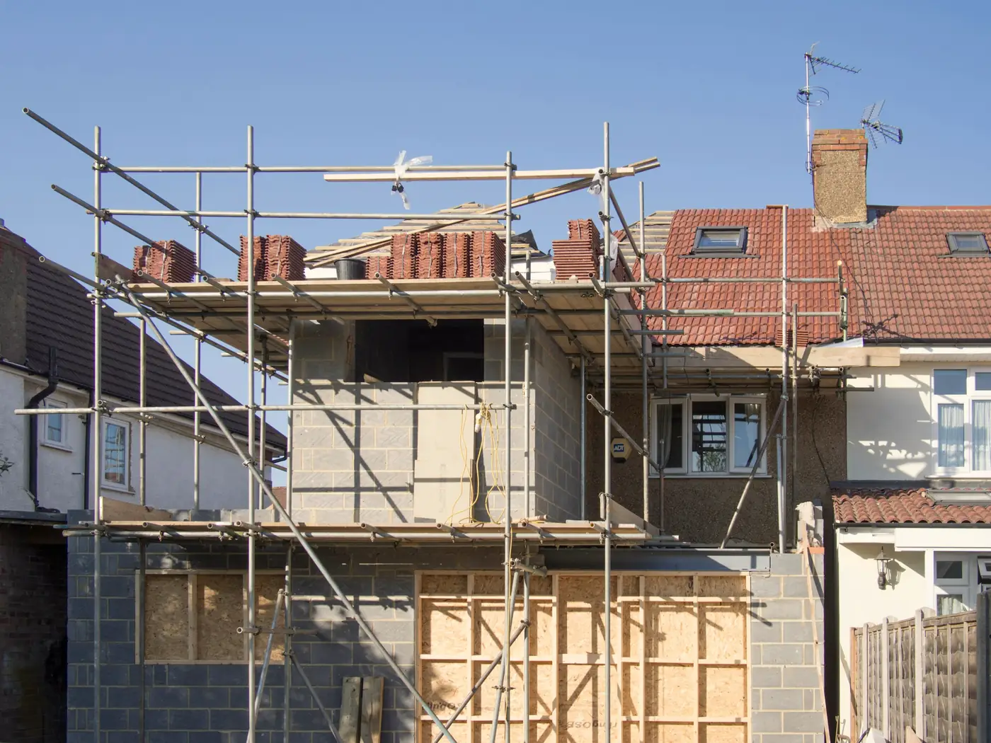 A two-storey rear extension in blockwork behind scaffolding, timber still stacked on the lawn