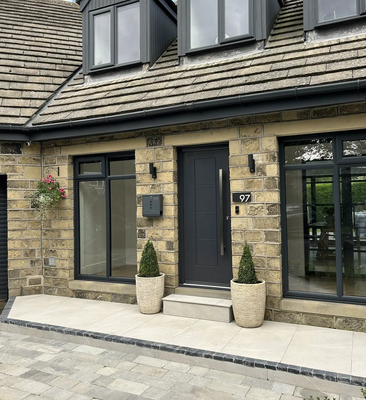 Front of a stone-faced house with slate-grey windows, a black front door and clipped bay trees